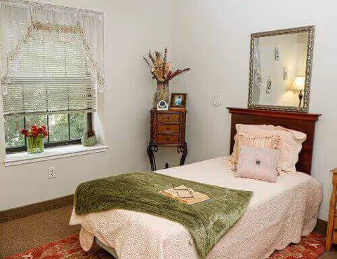 A cozy bedroom with a single bed covered in a light-colored bedspread and a green throw blanket. There are decorative pillows on the bed. A wooden nightstand with a framed photo and a vase with dried flowers is placed next to the bed. A large mirror hangs on the wall above the headboard. A window with blinds and a lace valance lets in natural light, and a small plant and flowers sit on the windowsill.