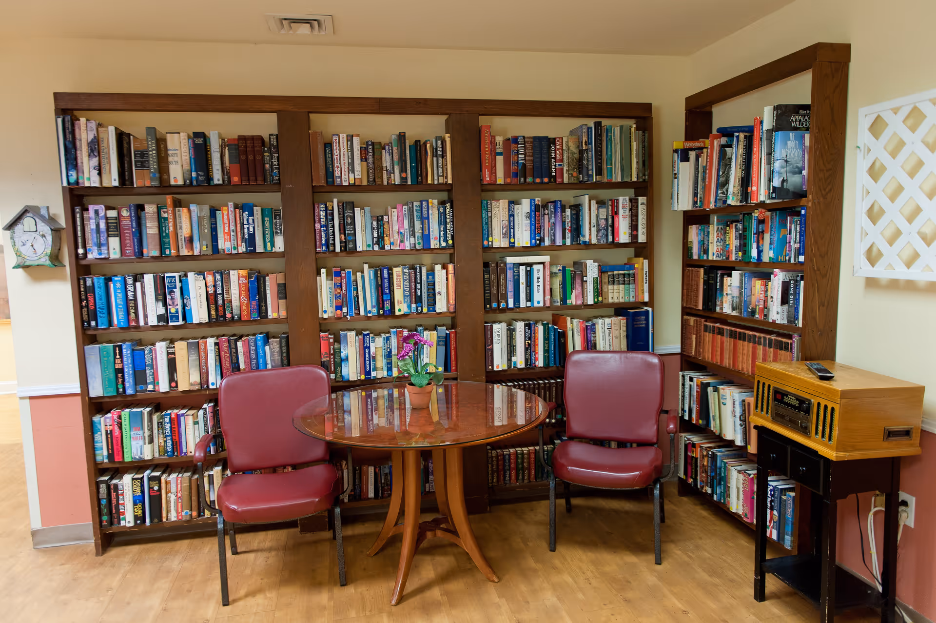 A cozy reading nook with wooden bookshelves filled with books, a round wooden table with a small potted plant, and two red cushioned chairs. There is a vintage-style radio on a small table to the right and a decorative wall clock on the left wall.