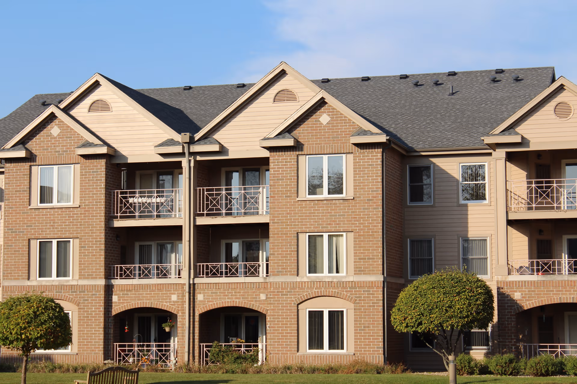Exterior view of a multi-story senior living facility building with brick and beige siding, featuring balconies and windows, with neatly trimmed trees and grass in the foreground under a clear blue sky.