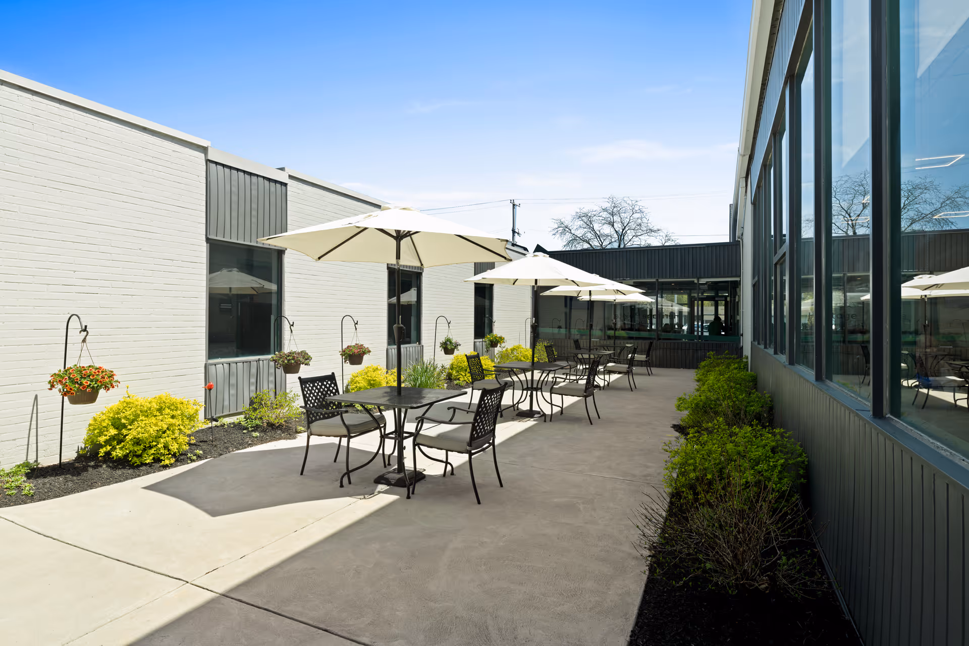 Outdoor patio area at Heritage Pointe Rehabilitation and Healthcare Center with several black metal tables and chairs, each shaded by large white umbrellas. The patio is surrounded by white and dark gray building walls, with windows on one side and hanging flower pots and shrubs along the other side. The sky is clear and blue.