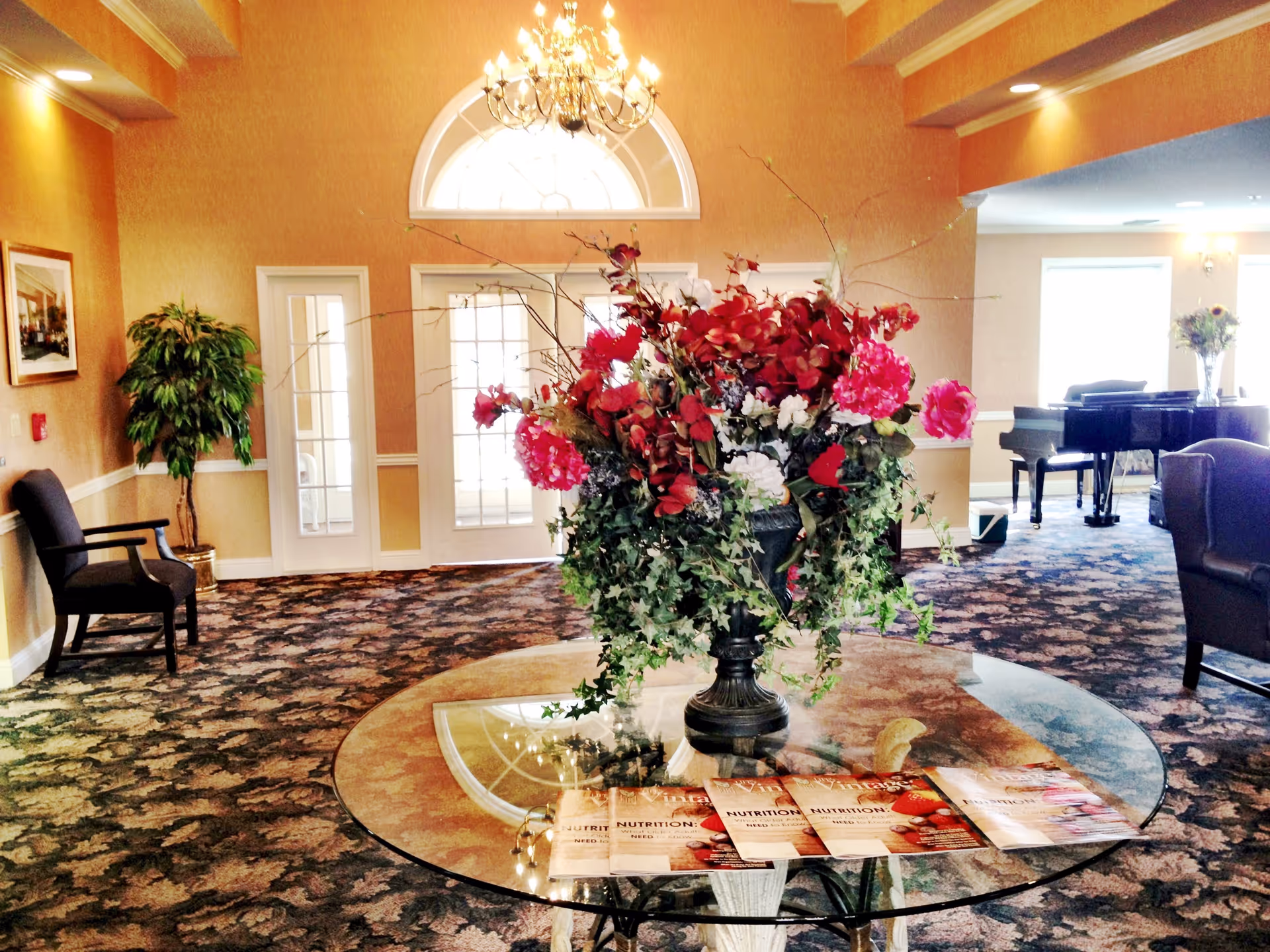 Interior view of a senior living facility common area with a round glass table in the foreground holding a large floral arrangement with red and pink flowers and greenery. The room has patterned carpet, beige walls, a chandelier, and large windows letting in natural light. There are chairs along the walls and a grand piano in the background.