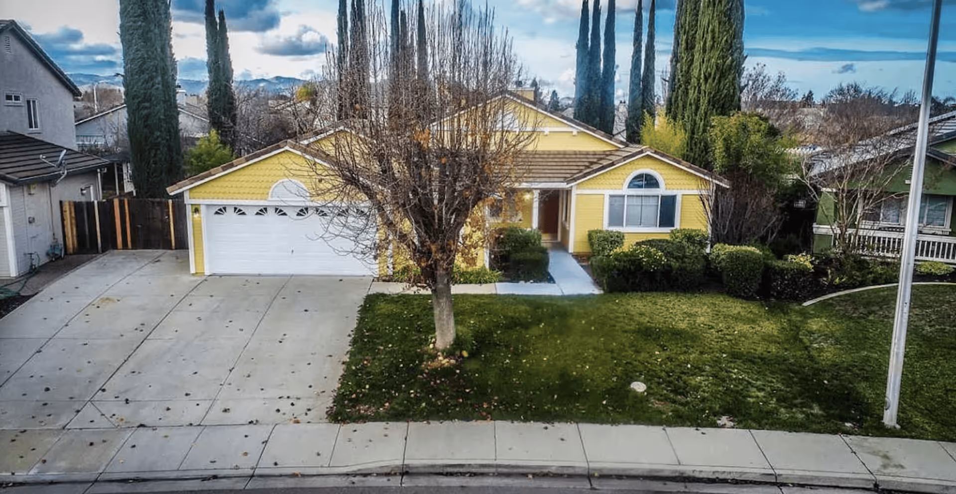 Front exterior view of a single-story yellow house with a white garage door, a leafless tree in the front yard, green bushes, and a well-maintained lawn. The house is situated in a suburban neighborhood with other houses and tall trees visible in the background under a partly cloudy sky.