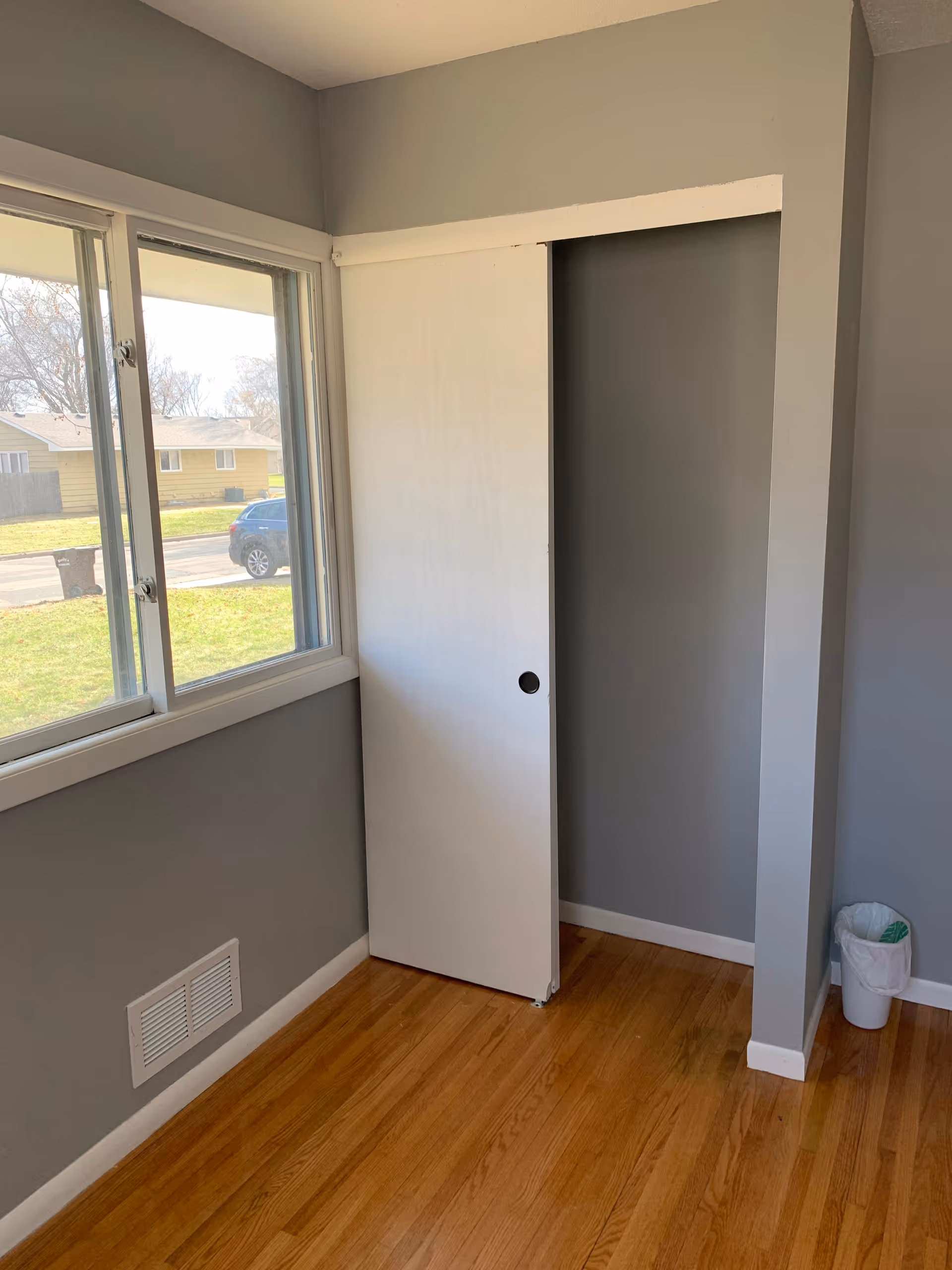 Empty room corner with gray walls, a wooden floor, a window showing a view of houses and a car outside, a white sliding closet door partially open, and a small trash can in the corner.