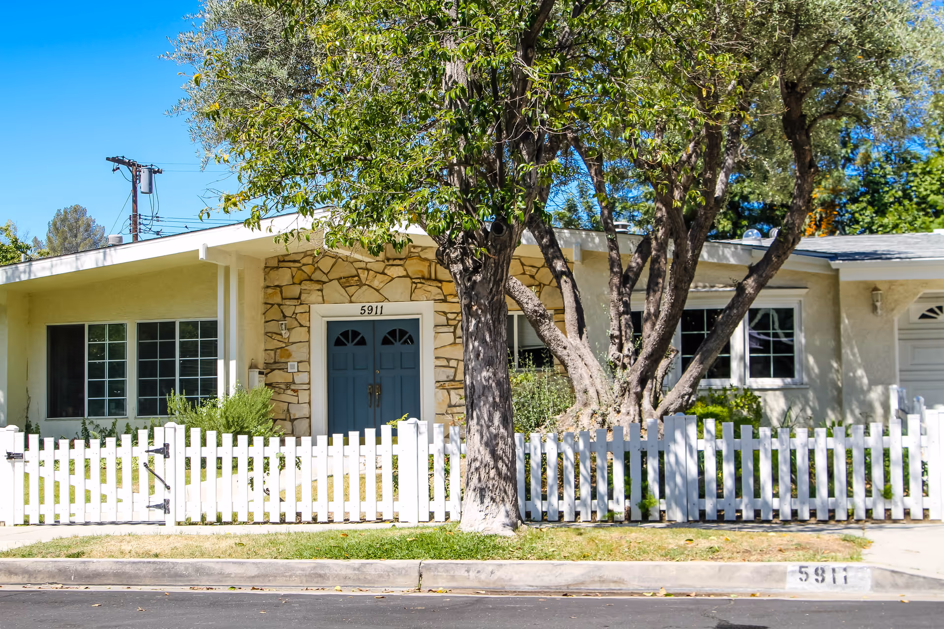 Single-story light-colored building with blue double front doors, a white picket fence, and large trees in front.