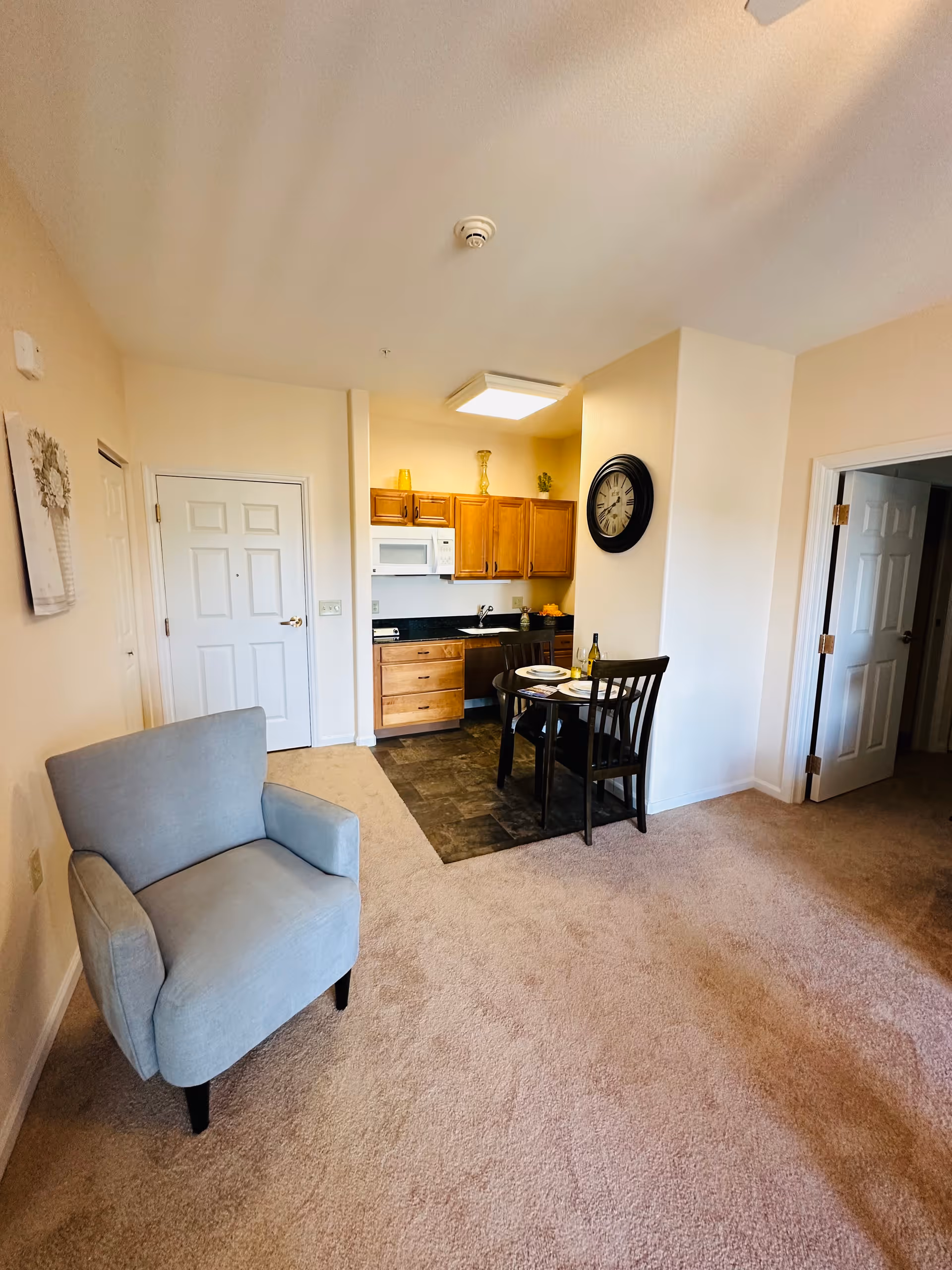 Open-plan living area with a light blue armchair, small dining table and a kitchenette with wooden cabinets and a wall clock.