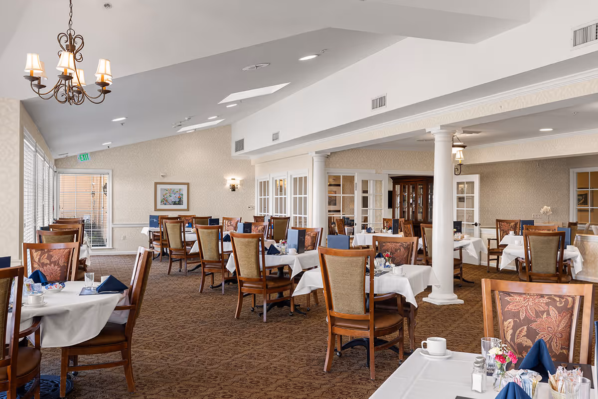 A spacious dining room with multiple tables covered in white tablecloths, each set with blue napkins, cups, and small flower arrangements. The room features wooden chairs with patterned upholstery, beige walls, carpeted floor, and large windows letting in natural light. Decorative columns and a chandelier add to the elegant ambiance.