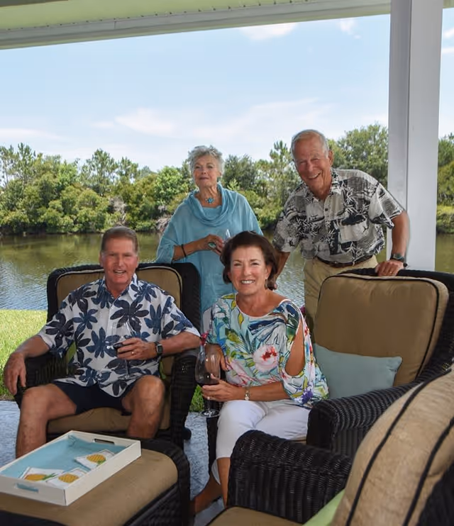 Four elderly adults sitting and standing on a covered patio overlooking a pond. Two men and two women are smiling and holding drinks, seated on wicker chairs with cushions. The background shows green trees and a blue sky with some clouds.