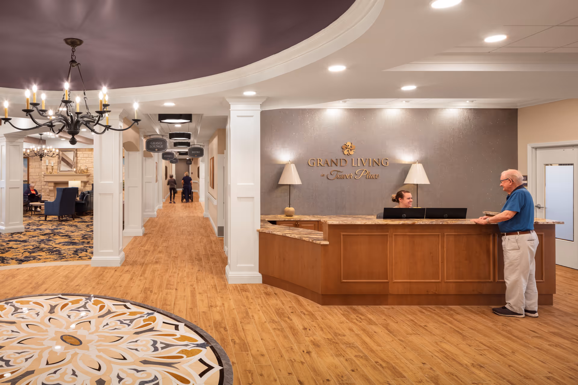 Reception area of Grand Living At Tower Place with a wooden front desk where a receptionist is smiling and talking to an elderly man. The floor has a decorative circular tile pattern near the entrance, and the ceiling features a chandelier. In the background, there is a hallway with signs indicating directions to the Grand Lounge, Studio, and Bistro, and a seating area with chairs and a fireplace.