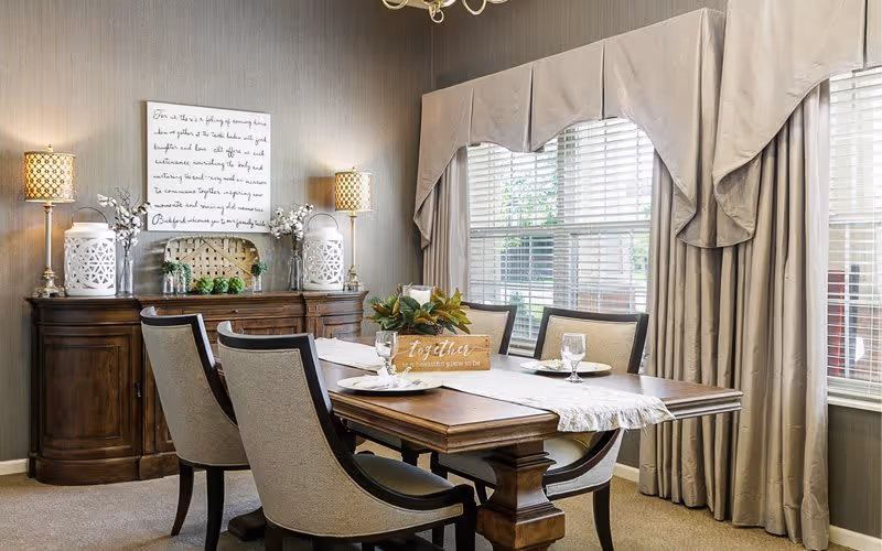 A well-lit dining room with a wooden dining table set for four, featuring beige cushioned chairs. The table has a white runner, glassware, and a decorative centerpiece with a sign that reads 'Together is a beautiful place to be.' Behind the table is a wooden sideboard with two lamps, decorative lanterns, and floral arrangements. Large windows with beige curtains allow natural light to fill the room.