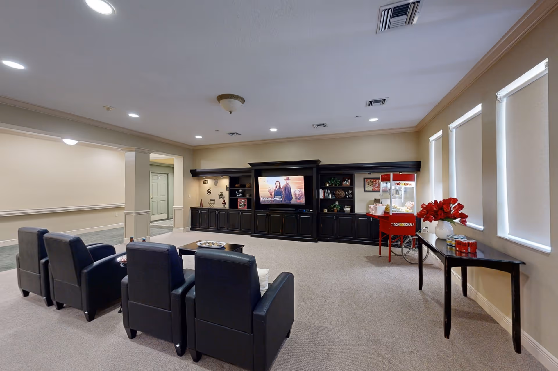 A cozy entertainment room with four black leather armchairs facing a large flat-screen TV mounted on a dark wooden cabinet. To the right, there is a red popcorn machine on a stand and a table with a vase of red flowers and some canned drinks. The room has beige walls, carpeted floor, and three windows with white blinds.
