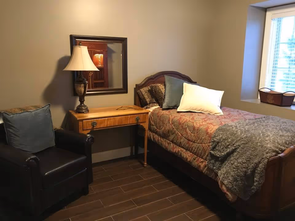 A cozy bedroom with a single wooden bed covered in a patterned bedspread, two pillows, and a gray throw blanket. Next to the bed is a wooden side table with a lamp and a mirror hanging on the wall above it. A dark leather armchair with a blue cushion is positioned beside the table. A window with blinds and a small basket on the windowsill is visible on the right side.