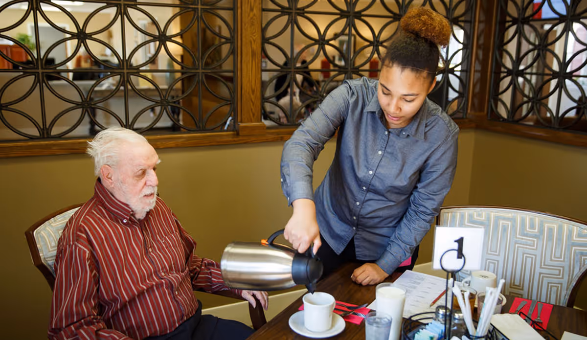 A young woman is pouring a beverage from a stainless steel carafe into a cup on a table while an elderly man in a red striped shirt sits nearby watching. They are seated in a dining area with decorative wooden lattice panels in the background.