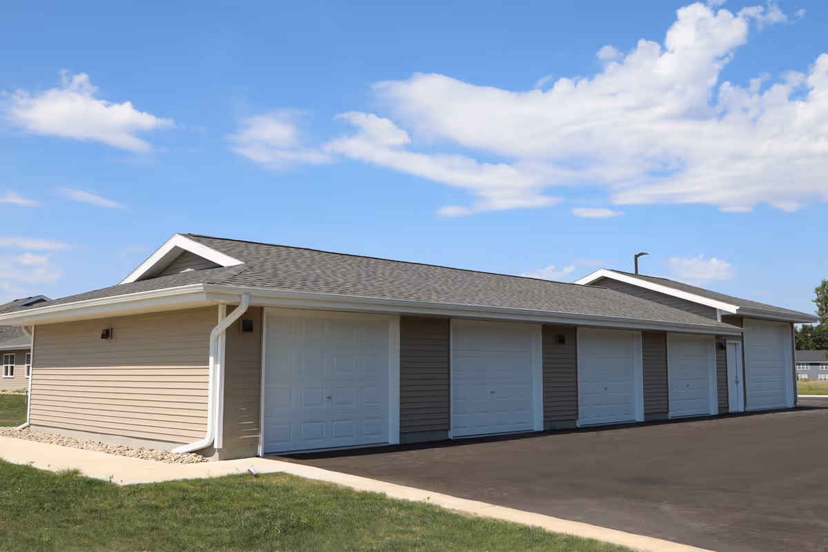 Exterior view of a beige building with multiple white garage doors under a partly cloudy blue sky, with a paved driveway and green grass surrounding it.