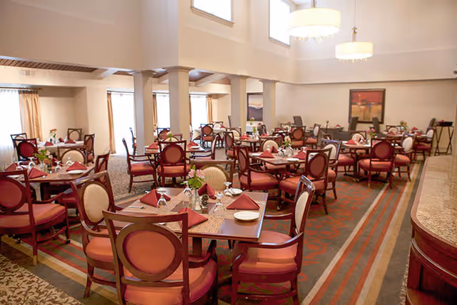 A spacious dining room with multiple wooden tables and red cushioned chairs arranged neatly. Each table is set with folded red napkins, plates, glasses, and small floral centerpieces. The room has large windows near the ceiling allowing natural light to brighten the space, and there are framed artworks on the walls.
