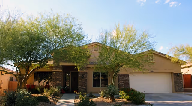 Front exterior view of a single-story assisted living facility building with a beige facade, stone accents around the entrance and garage, two trees, and desert landscaping with shrubs and rocks under a clear blue sky.