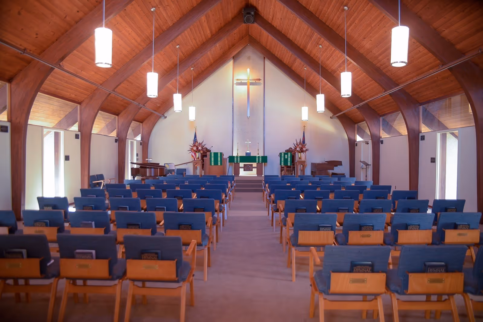 Interior view of a chapel with wooden vaulted ceiling and hanging lights. Rows of blue cushioned chairs with hymnals are arranged facing an altar with a cross on the wall behind it. There are flower arrangements and musical instruments on either side of the altar.