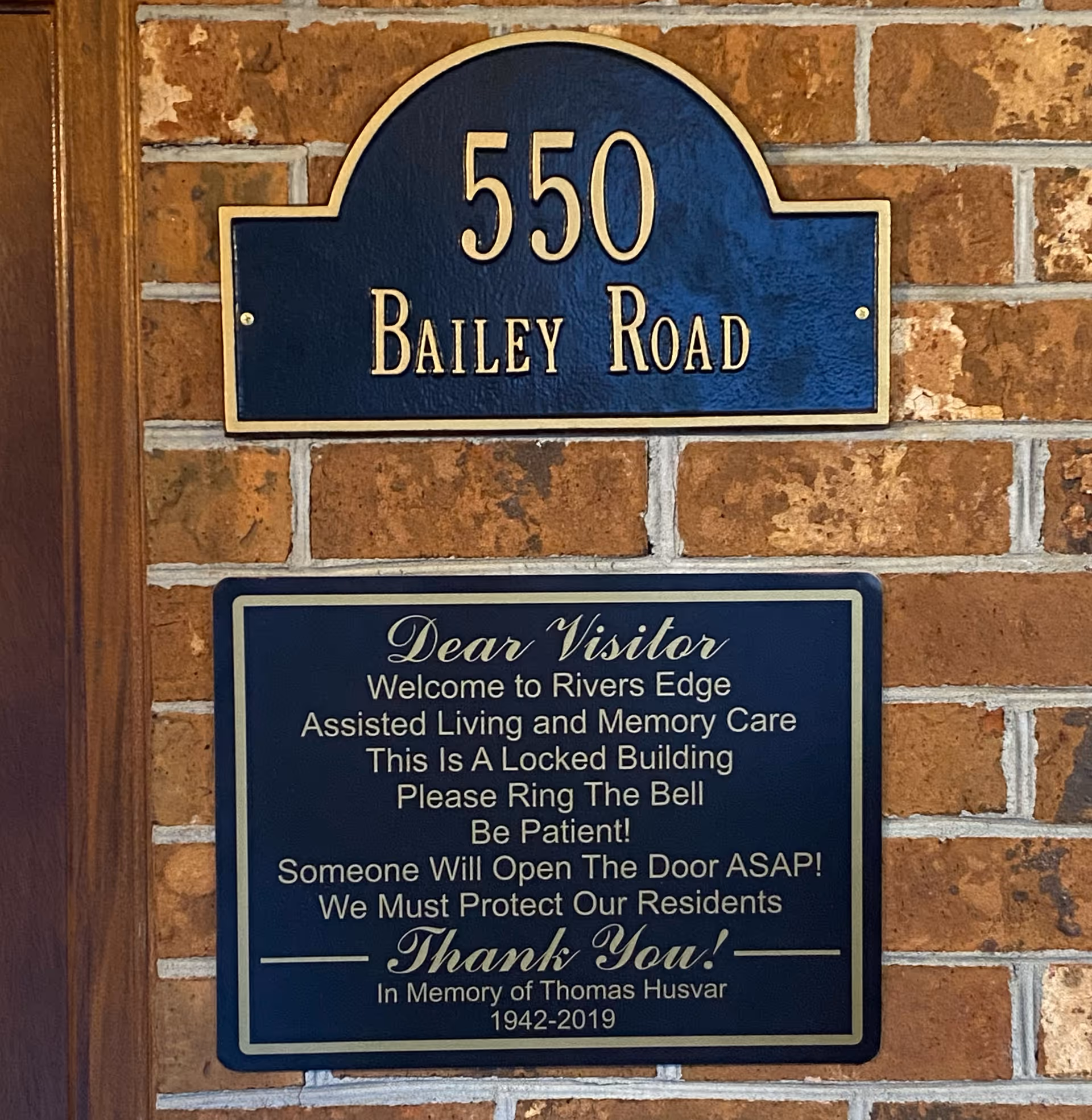 Two plaques mounted on a brick wall. The top plaque is blue with gold trim and text displaying the address '550 Bailey Road'. The bottom plaque welcomes visitors to Rivers Edge Assisted Living and Memory Care, instructing them to ring the bell as the building is locked, and asks for patience while someone opens the door. It also includes a memorial note for Thomas Husvar, 1942-2019.