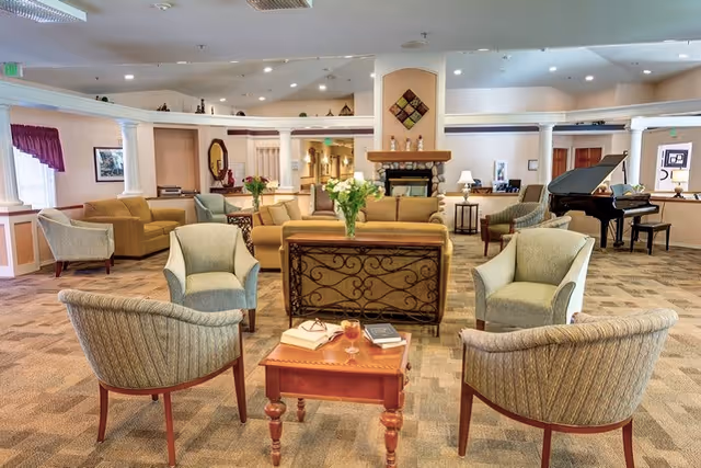 A spacious and well-lit living room area in a senior living facility with multiple armchairs and sofas arranged around a small wooden table with books and a drink. There is a fireplace in the background, a grand piano to the right, and decorative elements such as flowers and framed pictures on the walls.