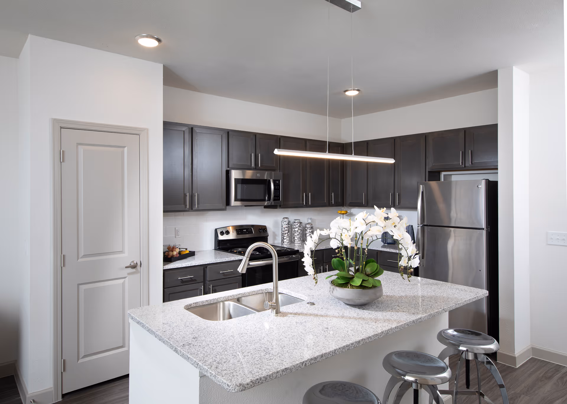 Modern kitchen with dark wood cabinets, stainless steel appliances including a refrigerator, microwave, and stove. A large granite island with a built-in sink and a silver faucet is in the center, decorated with a white orchid plant. Three metal bar stools are positioned at the island. The walls are white, and the floor has a wood finish.
