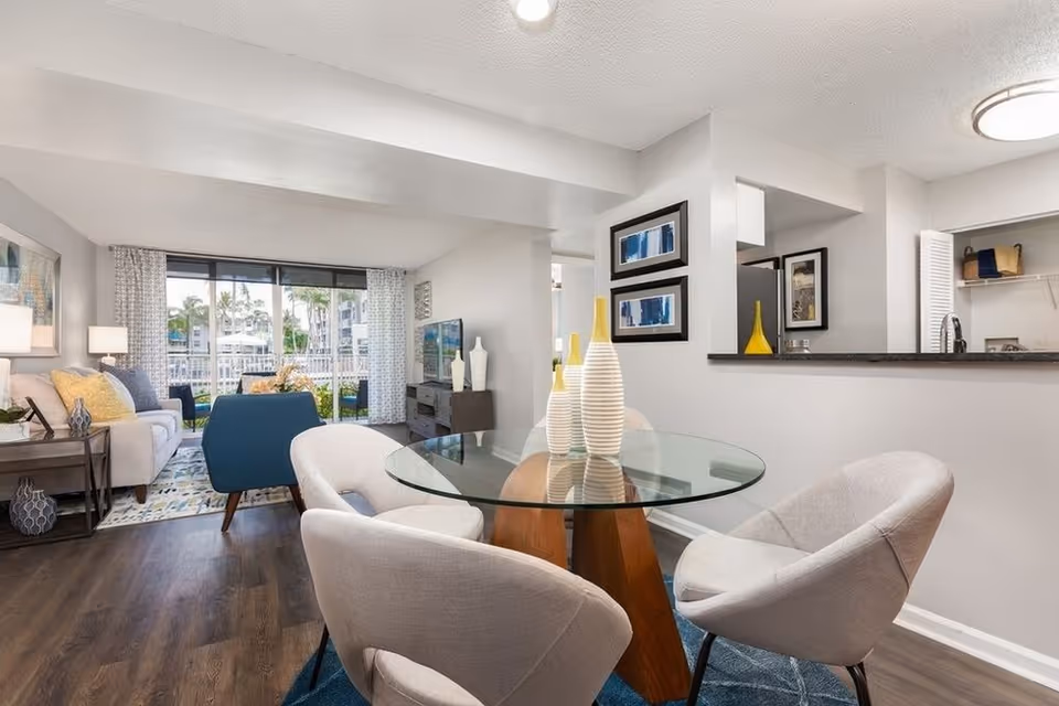 A modern living and dining area with a round glass dining table surrounded by four light gray upholstered chairs. The living room features a white sofa with yellow and gray pillows, a blue armchair, a TV on a dark wooden stand, and large windows with sheer curtains overlooking an outdoor area with palm trees. The walls are light gray, and the floor is dark wood. Decorative vases and framed artwork add accents to the space.