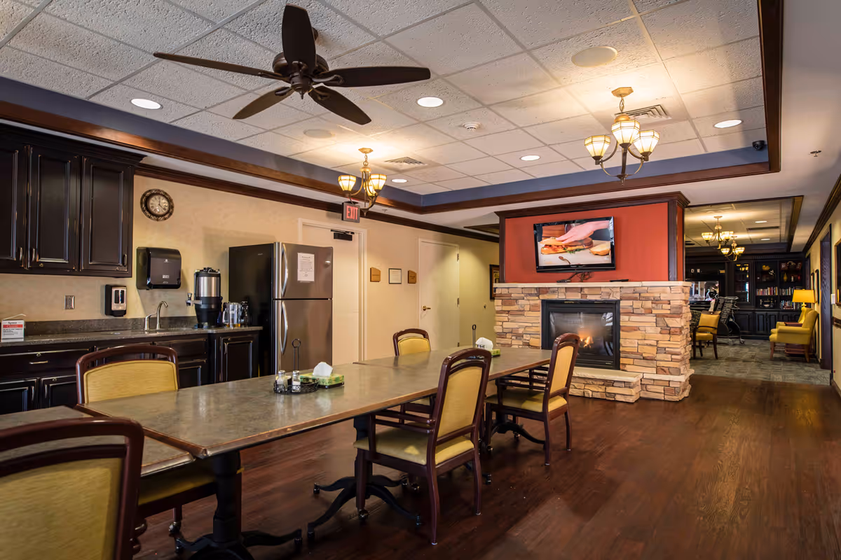 A cozy common area in Pioneer Ridge Independent Living of Lawrence featuring a long table with chairs, a stone fireplace with a TV mounted above it, dark wood cabinetry with a countertop, a refrigerator, and ceiling fans and lights. The room has warm lighting and wood flooring, with a hallway leading to another seating area with yellow chairs and bookshelves.