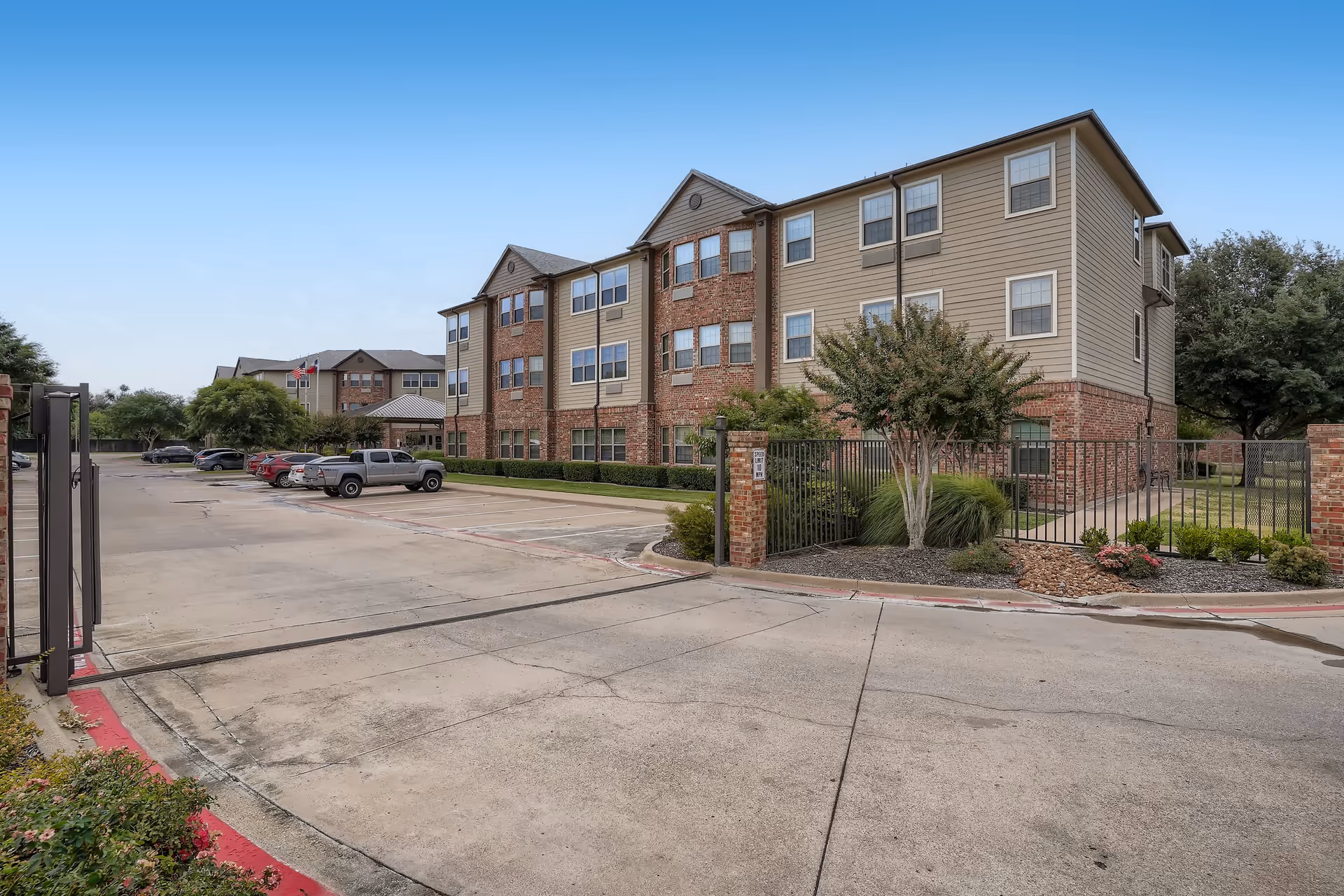 Exterior view of a three-story senior living facility building with a combination of brick and siding. There is a gated entrance with landscaping including bushes and small trees. Several cars are parked in the parking lot adjacent to the building under a clear blue sky.