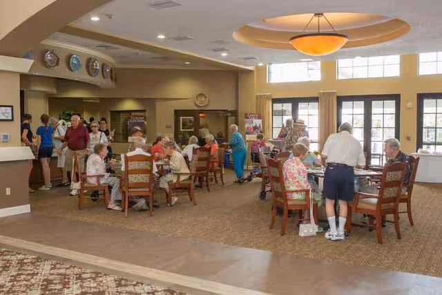 A bright communal dining room in a senior living facility with residents seated at tables socializing and staff in the background.
