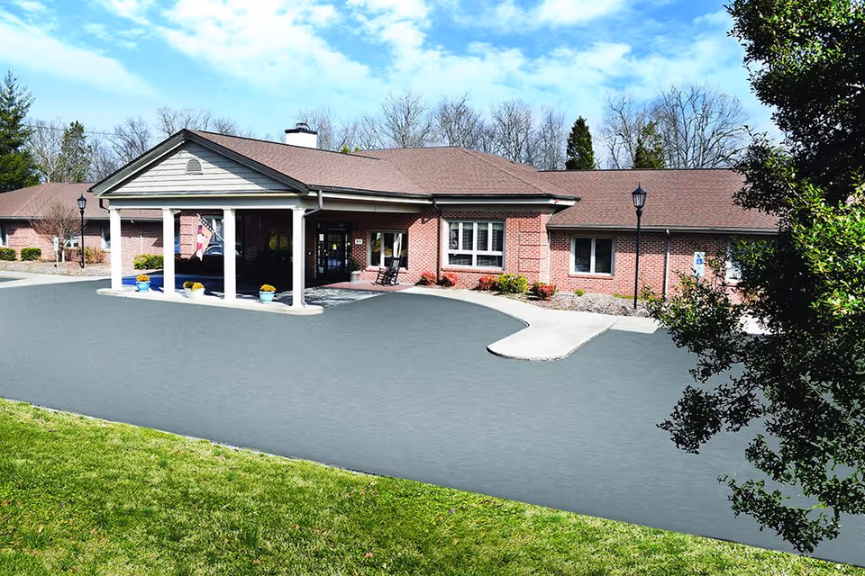 Exterior front view of a single-story brick building with a covered entrance supported by white columns. There are potted plants near the entrance, a flag, and a paved driveway leading up to the building. Trees and grass surround the area under a partly cloudy sky.