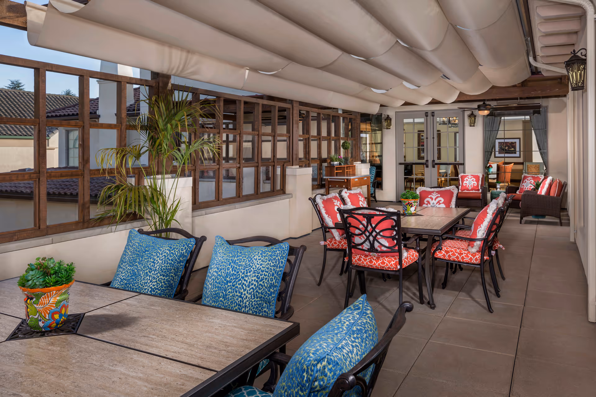 Covered outdoor patio area with multiple tables and chairs featuring colorful cushions in blue and red patterns. Potted plants are placed on the tables and near the wooden lattice windows. The space is well-lit with wall-mounted lantern-style lights and ceiling fans, leading to an indoor area visible through glass doors.