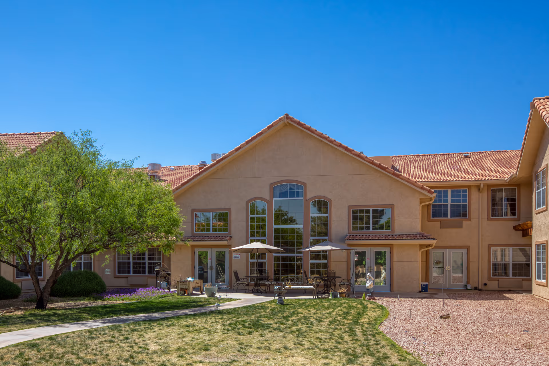 Exterior view of Vista Pointe at Sierra Vista senior living facility showing a beige stucco building with large windows, a tiled roof, and a courtyard with patio tables, umbrellas, chairs, a tree, and a grassy area under a clear blue sky.