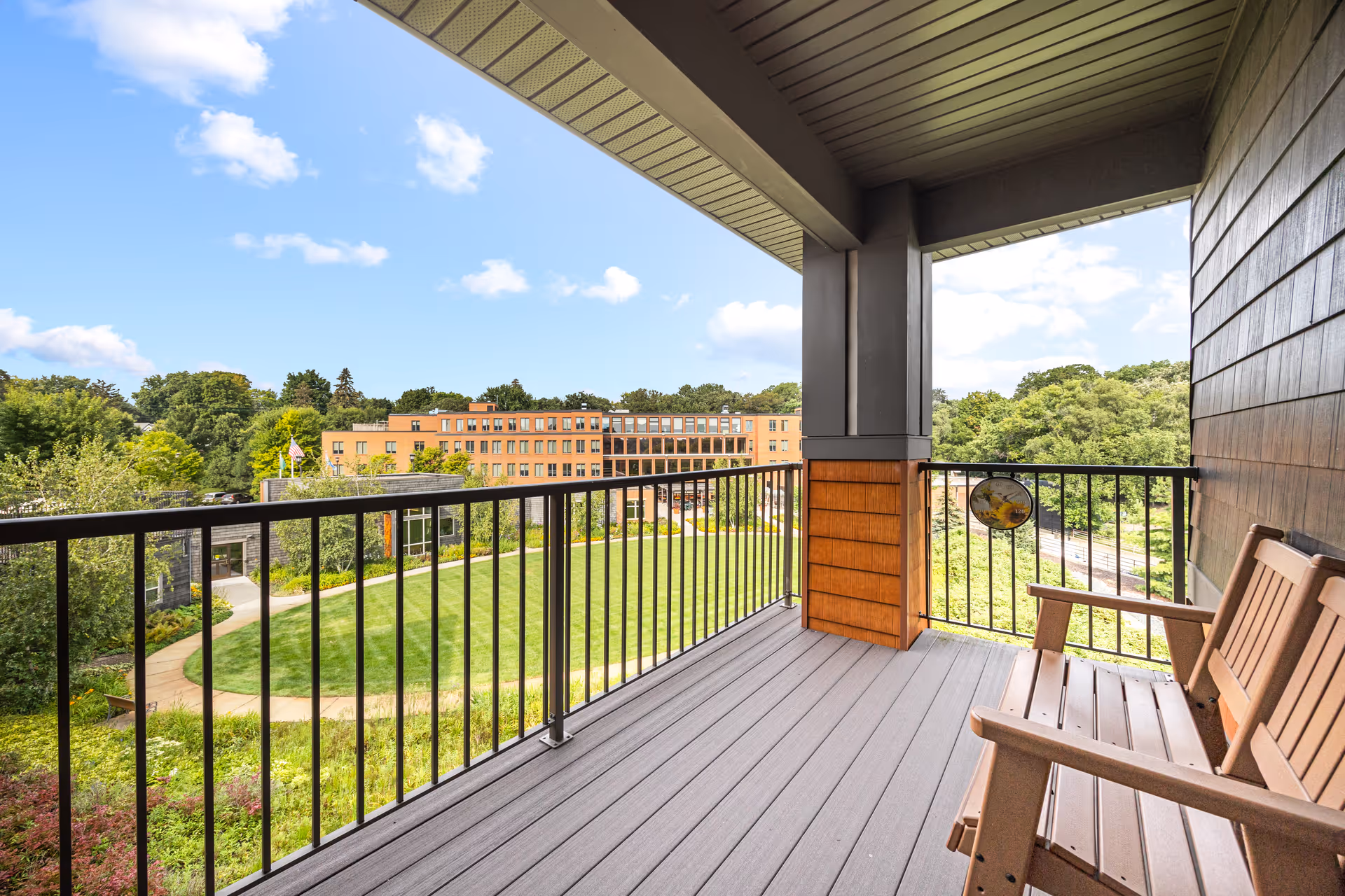 Balcony with wooden chairs overlooking a landscaped courtyard and a multi-story building under a blue sky.