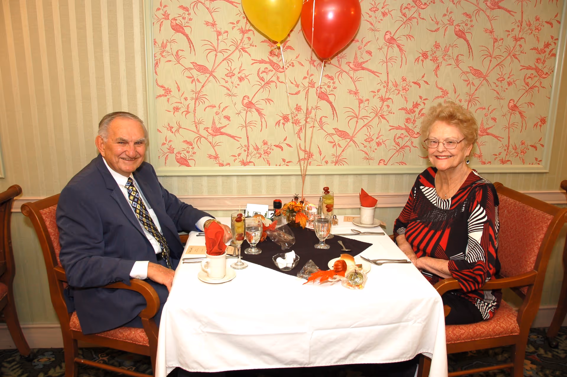 An elderly man and woman sitting at a dining table in a decorated room. The man is wearing a suit and tie, and the woman is wearing a patterned dress. The table is set with glasses of water, cups, utensils, a small centerpiece with autumn decorations, and two balloons (one yellow and one red) tied to the centerpiece. The background features wallpaper with a bird and floral pattern.