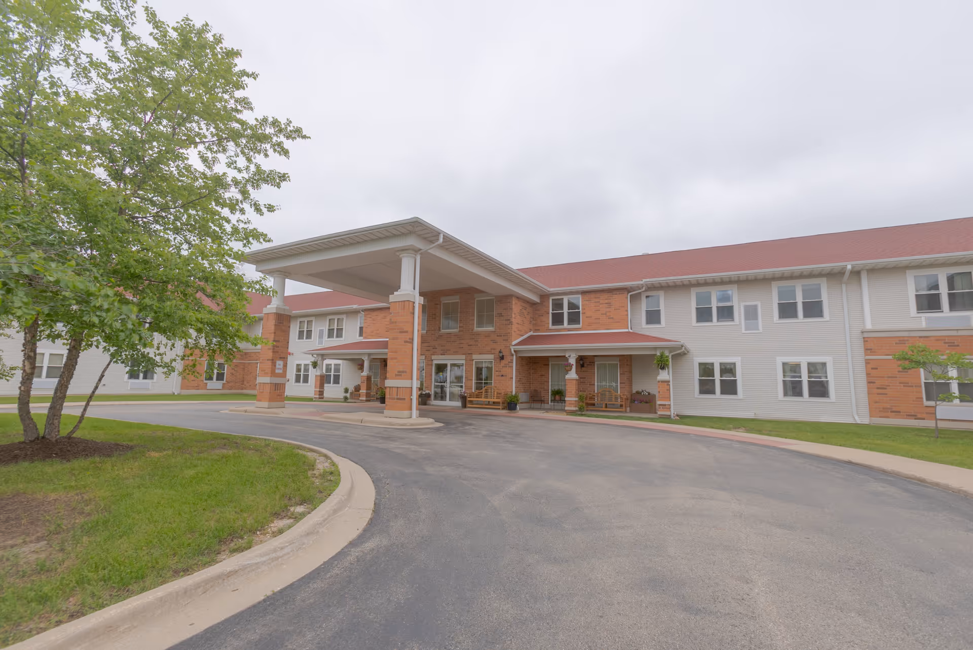 Front exterior of a two-story senior living facility with a covered entrance, circular driveway, lawn and trees.