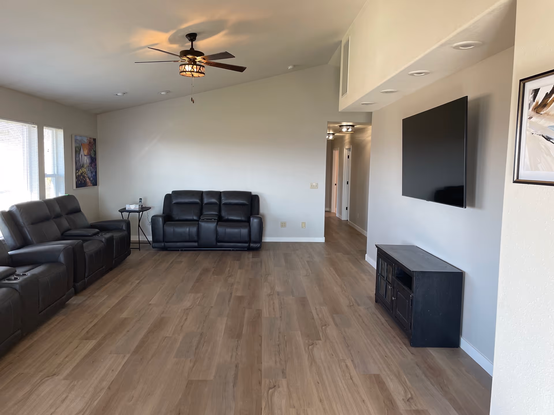 A spacious living room with light wood flooring, two black leather sofas, a small side table with a phone, a ceiling fan with a light fixture, a wall-mounted flat screen TV, and a black TV stand. There is a hallway visible in the background and a framed abstract painting on the wall.