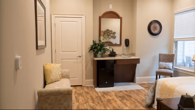 A cozy assisted living room featuring a small beige loveseat with yellow and beige pillows on the left, a wooden floor, a white door, a small sink with a mirror above it, a plant on the sink counter, a wall clock, a cushioned chair near a window with blinds, and part of a bed with a beige and white comforter on the right.
