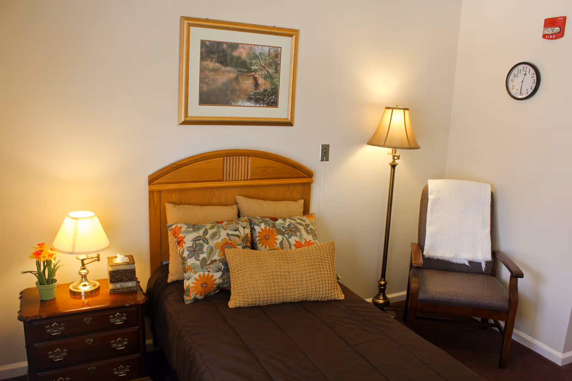 A cozy bedroom with a wooden headboard bed adorned with multiple floral and textured pillows. There is a wooden nightstand with a lamp, a small plant, and a decorative box. A floor lamp stands next to a cushioned armchair with a white throw draped over it. A framed nature painting hangs above the bed, and a clock is mounted on the wall.