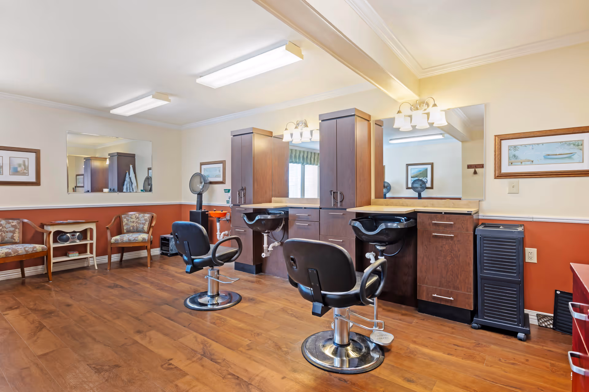 Interior view of a hair salon area in a senior living facility with two black salon chairs in front of wooden cabinets and sinks. The room has wood flooring, framed pictures on the walls, a large mirror, and two upholstered chairs along one wall.