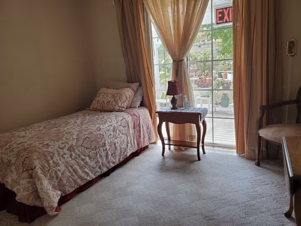 A cozy bedroom with a single bed covered in a floral patterned bedspread and matching pillows. Next to the bed is a small wooden side table with a red lamp and a bottle of hand sanitizer. The room has beige carpet and beige walls. Sheer curtains frame a sliding glass door that leads to an outdoor patio with plants visible outside. An exit sign is visible above the door.