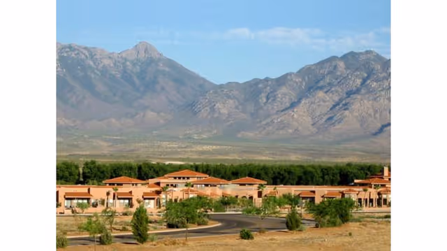 View of a senior living facility named Silver Springs with multiple single-story buildings surrounded by trees and landscaping, set against a backdrop of large rocky mountains under a blue sky.