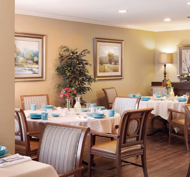 A dining room with round tables covered in white tablecloths, set with turquoise plates, cups, and silverware. Wooden chairs with striped cushions surround the tables. The room has beige walls adorned with framed landscape paintings and a potted plant. A lamp on a side table provides warm lighting.