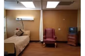 Nursing facility room with a hospital bed, privacy curtain, an upholstered armchair, and a small TV on a stand.