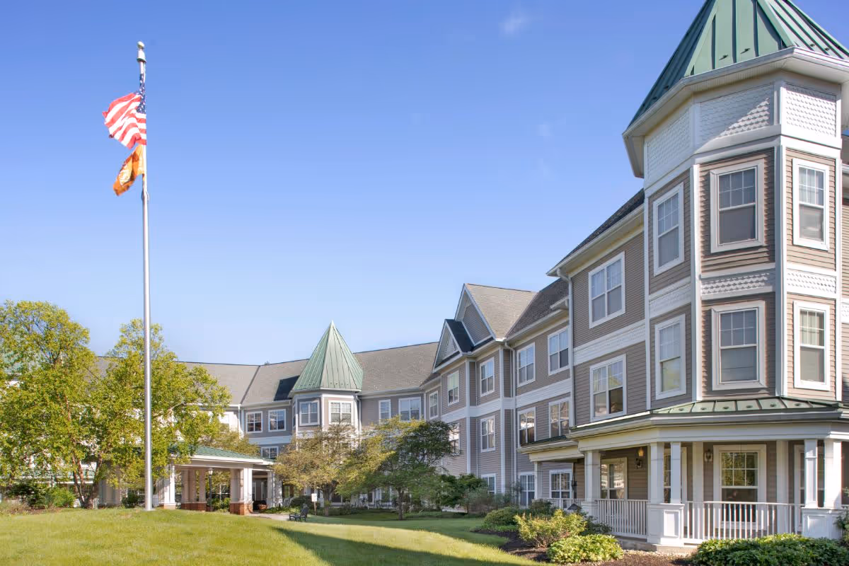 Exterior view of a large senior living facility building with multiple windows and a green roof. There is a well-maintained lawn with trees and shrubs in front, and two flags flying on a flagpole against a clear blue sky.