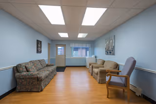 A small common area with light blue walls and a wood floor. The room contains a floral patterned sofa on the left, two beige armchairs in the middle, and a wooden chair with purple upholstery on the right. There is a window with a valance and a door at the far end of the room. Two framed pictures hang on the walls.