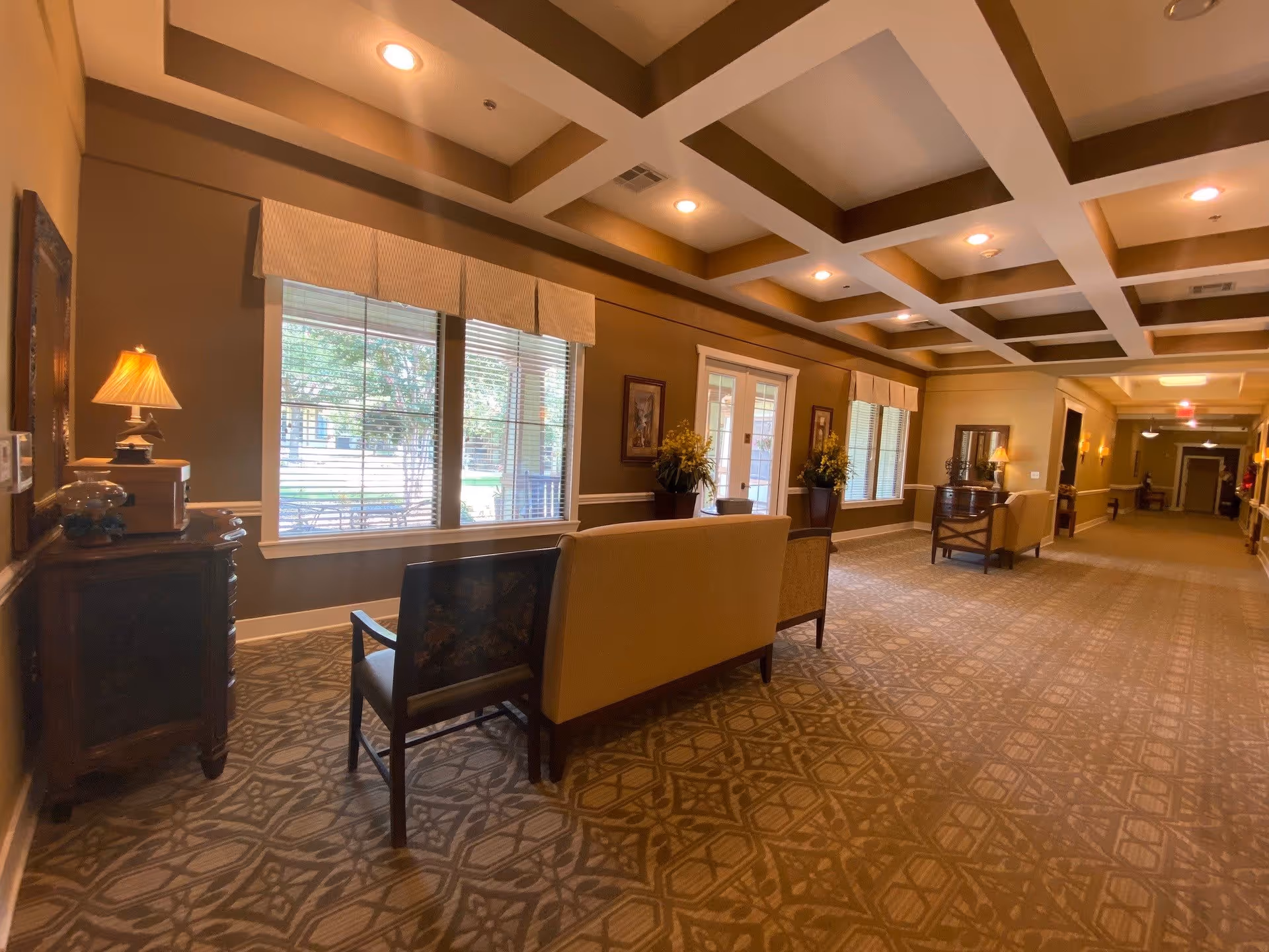 A spacious, well-lit hallway in an assisted living facility with patterned carpet flooring, beige walls, and a coffered ceiling with recessed lighting. The hallway features seating areas with chairs and a sofa, decorative plants, framed artwork on the walls, and large windows with valances letting in natural light.