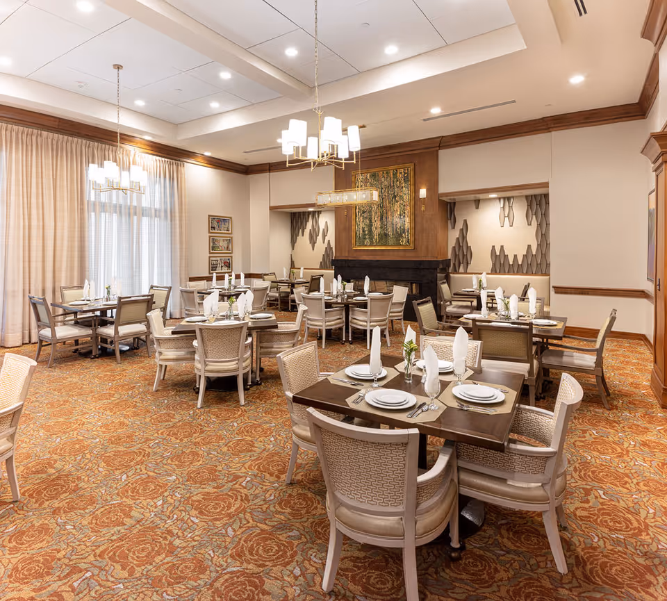 Elegant dining room with multiple tables set with white plates, napkins, and silverware. The room features patterned carpet, beige upholstered chairs, large windows with beige curtains, and modern chandeliers hanging from a white ceiling. There is a decorative fireplace with artwork above it and textured wall panels in the background.