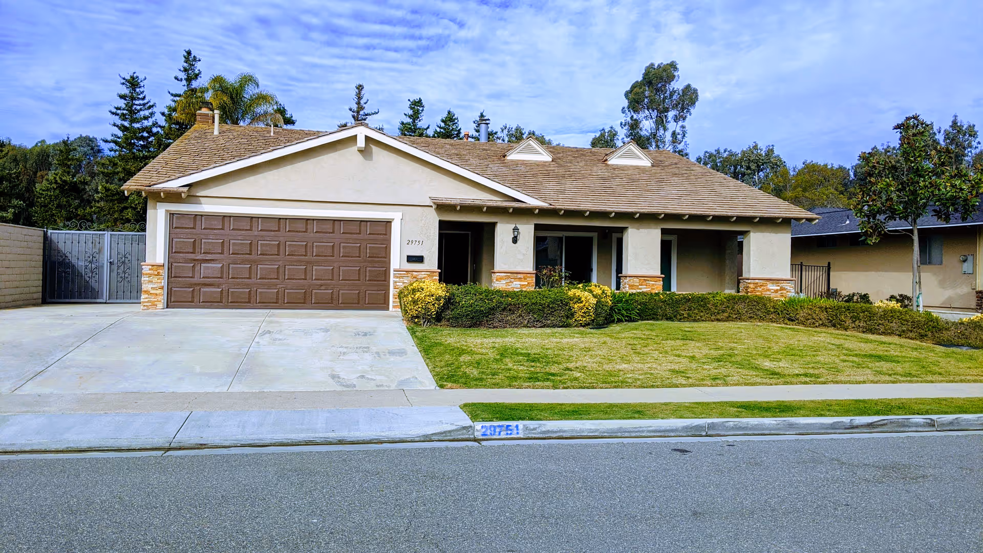 Single-story suburban house with a two-car garage, front lawn, driveway, and shrubs under a partly cloudy sky.