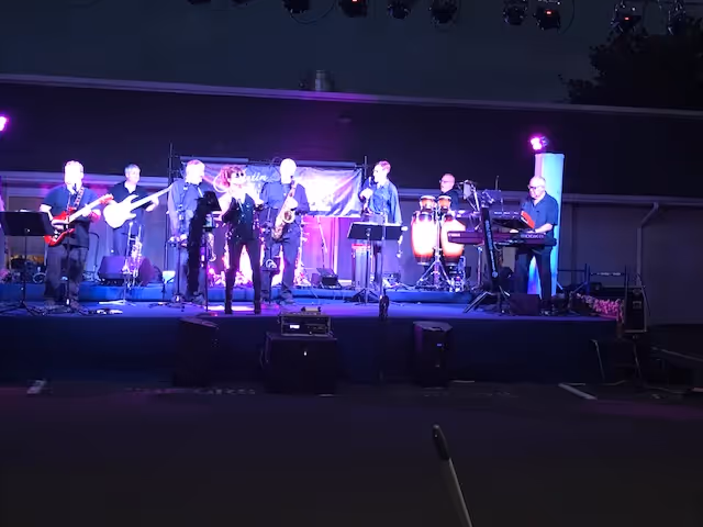 A band performing on an outdoor stage at night with purple lighting. The band members are playing various instruments including guitars, saxophone, keyboard, and percussion.