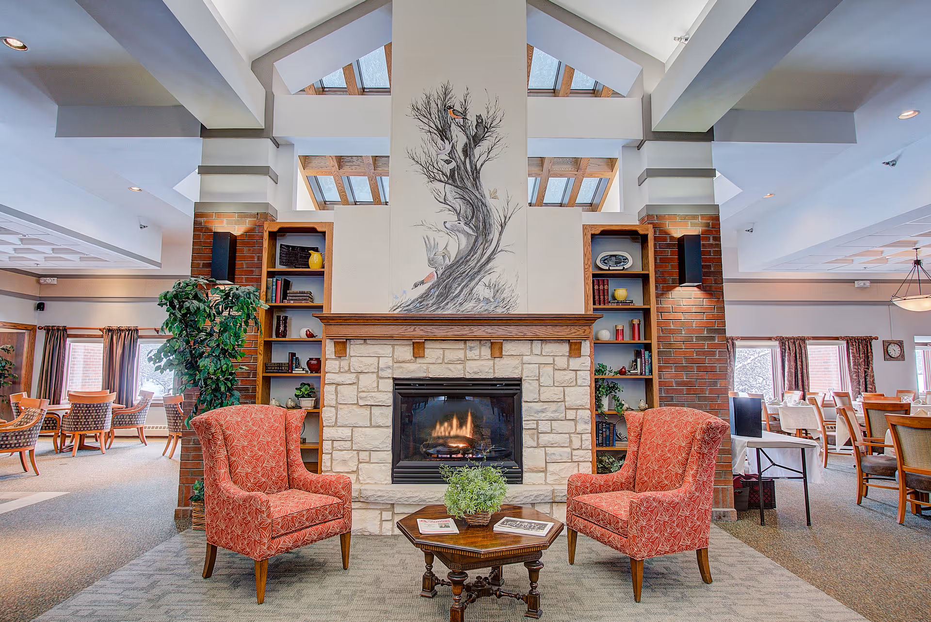 A cozy senior living community common area featuring a stone fireplace with a nature-themed mural above it. Two red patterned armchairs face a small wooden coffee table with a plant and magazines. Built-in shelves flank the fireplace, holding books and decorative items. Large windows with curtains allow natural light into the spacious room, which includes additional seating and dining tables in the background.