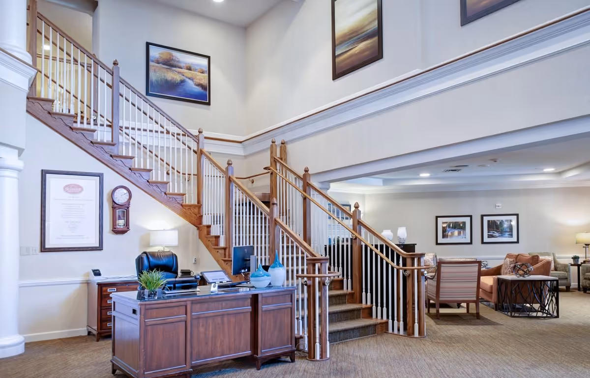Interior view of a senior living facility lobby with a wooden reception desk, a black office chair, and a computer monitor. Behind the desk is a staircase with wooden handrails and white balusters leading to an upper floor. The walls are decorated with framed landscape paintings and a wall clock. To the right, there is a seating area with chairs, a sofa, side tables, and lamps.