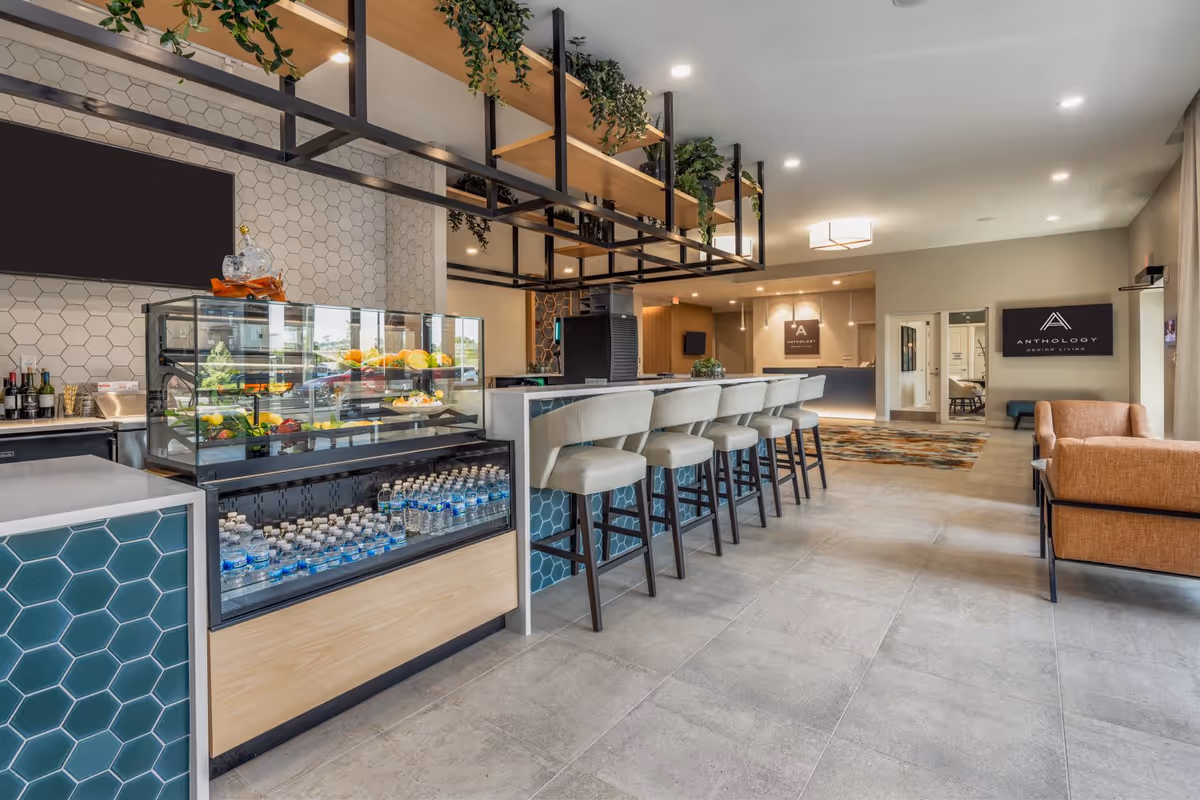 Interior view of a modern senior living facility lounge area with a long counter and bar stools, a display case with bottled water and fruit, hanging shelves with plants, and a seating area with orange armchairs. The reception desk and signage for Anthology Senior Living are visible in the background.